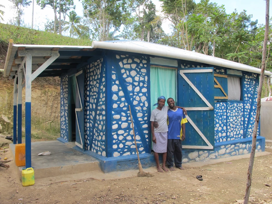 Two smiling people in front of a small blue and white house. The solid dwelling, built with local materials, offers protection and security.