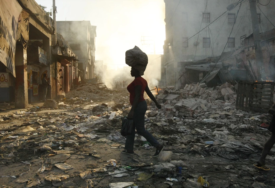 A woman walks through a destroyed town in Haiti carrying a bag on her head at sunset after the 2010 earthquake supported by Swiss Solidarity’s relief efforts.