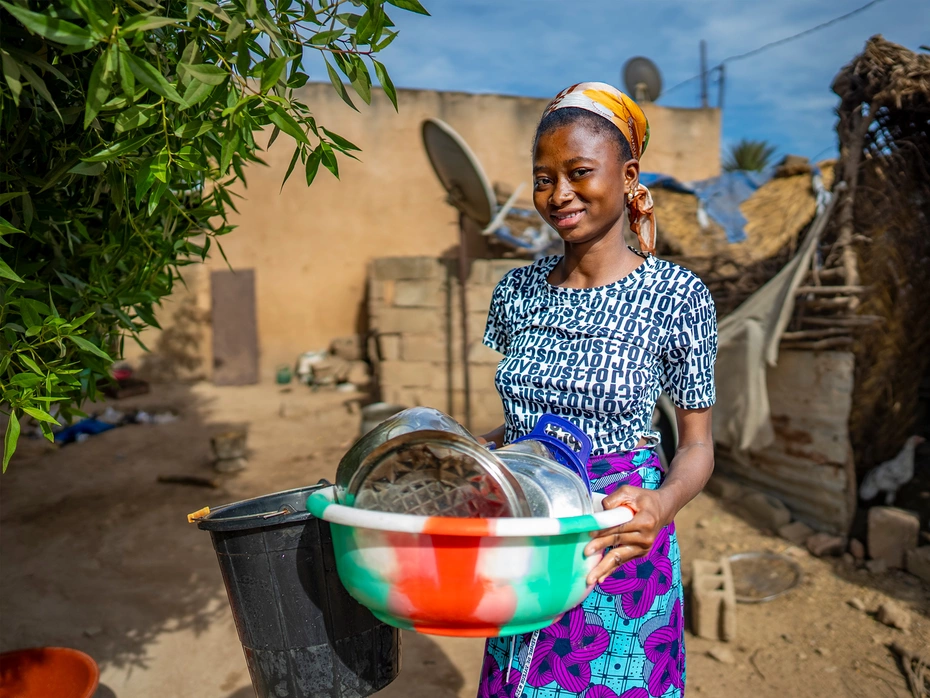 A woman smiles while carrying a large basket full of dishes in front of a hut.