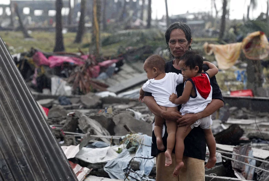 A man smiling tightly while holding two children amidst destruction caused by Typhoon Haiyan in 2013, showing resilience and hope.