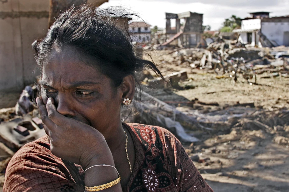A woman with tears in her eyes looks to the side, standing in front of a devastated landscape after the 2004 tsunami in Southeast Asia.