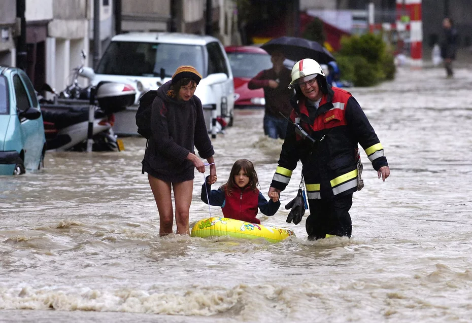 A man and woman holding the hands of a child sitting on a yellow inflatable boat walking through flooded streets during the 2005 Swiss floods relief efforts.