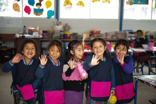 Five young girls dressed in blue and pink are happily smiling and waving at the camera in their classroom.